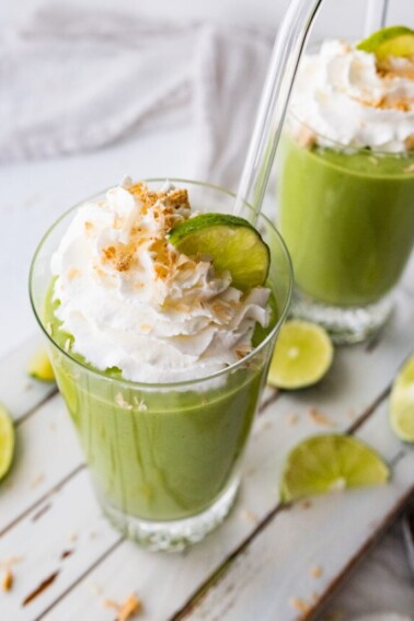 Two key lime pie smoothies in glasses with straws topped with whip cream, toasted coconut and lime slices.