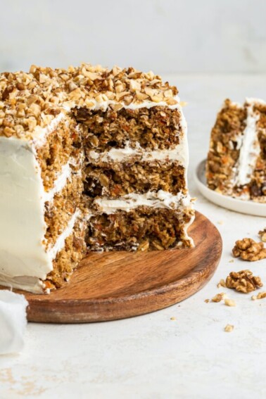 3-layer oatmeal carrot cake, frosted with chopped walnuts on top served on a wooden board. A few slices of the cake have been cut out exposing the inside of the cake.