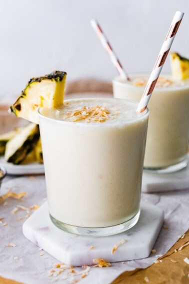 Two pineapple smoothies with straws, topped with toasted coconut and fresh pineapple slices.