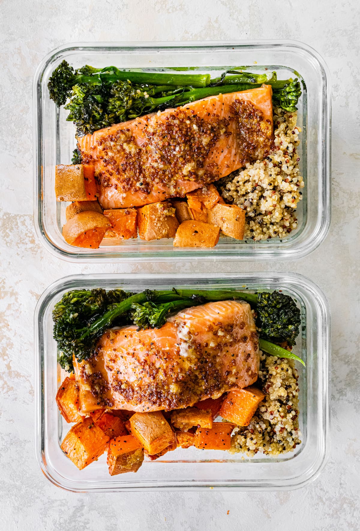 Two glass storage containers with a bed of quinoa topped with roasted sweet potatoes and broccolini and mustard maple salmon.
