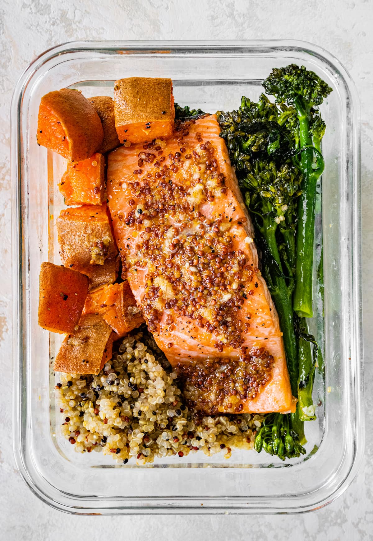 One glass storage container with a bed of quinoa topped with roasted sweet potatoes and broccolini and mustard maple salmon.