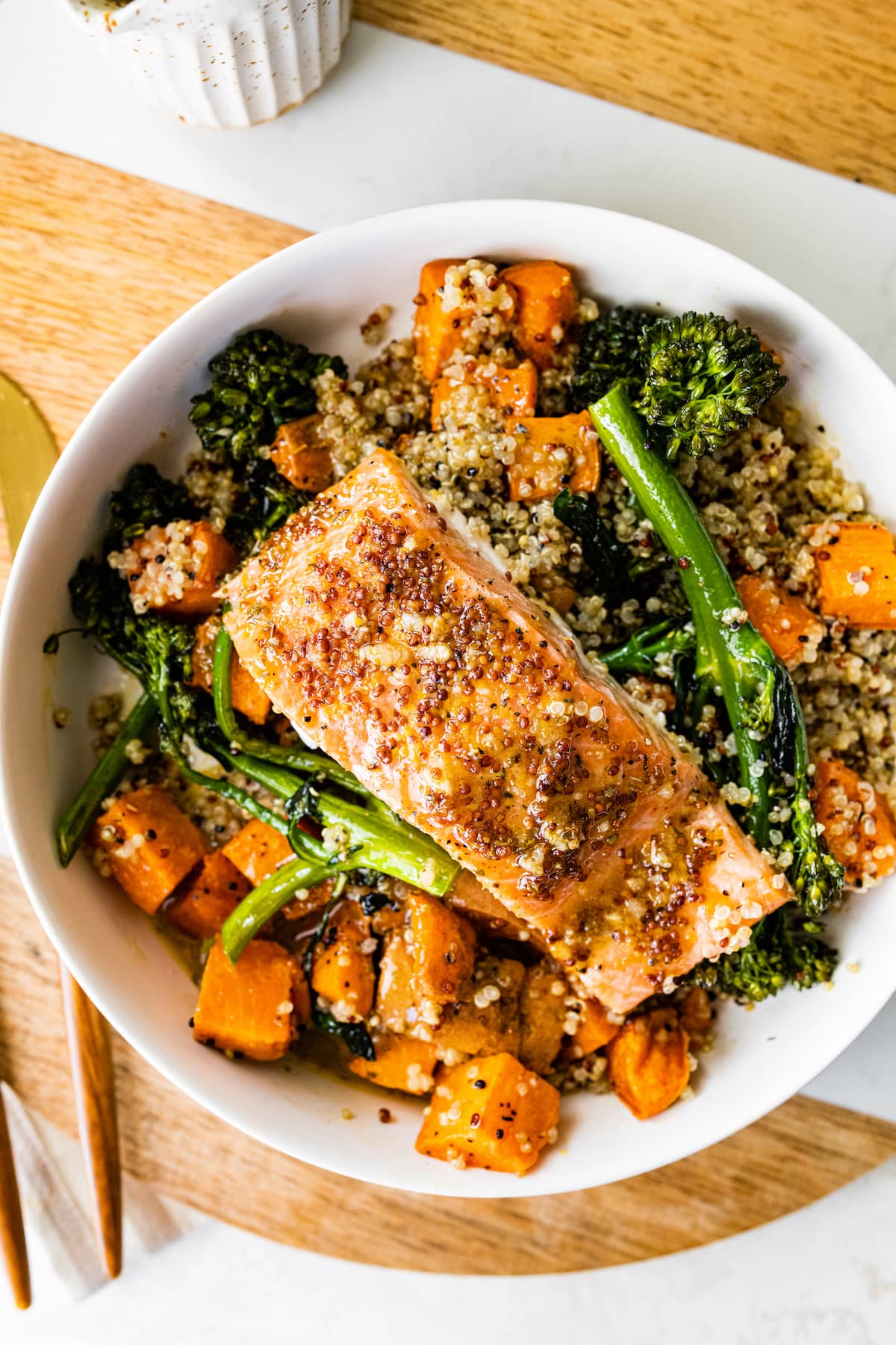 A white serving bowl with tri-colored quinoa and roasted sweet potatoes and broccolini mixed together and topped with mustard maple salmon.