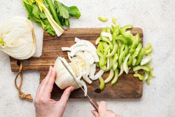 Woman's hands slicing fennel and celery on a wooden cutting board.