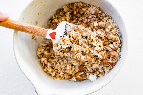 Spatula mixing oats, almonds and shredded coconut in a mixing bowl.