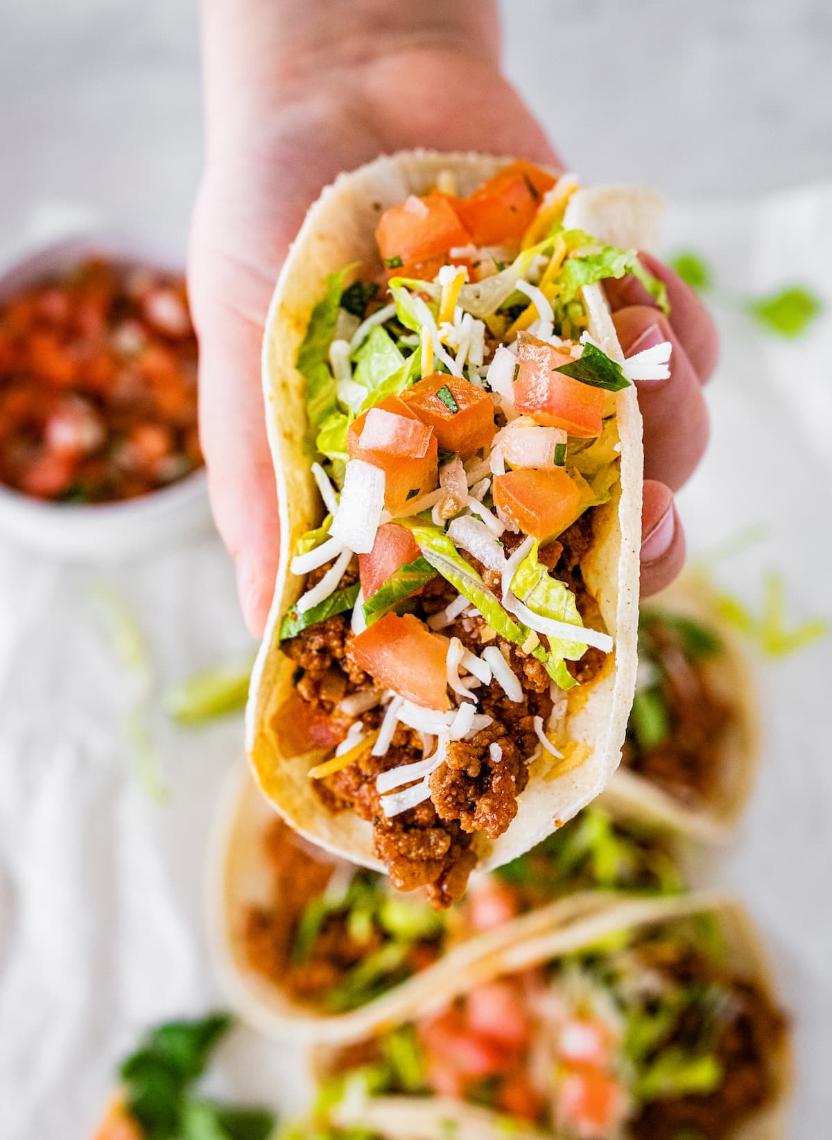 Woman's hand holding one ground turkey taco topped with lettuce, tomatoes, shredded cheese and cilantro.