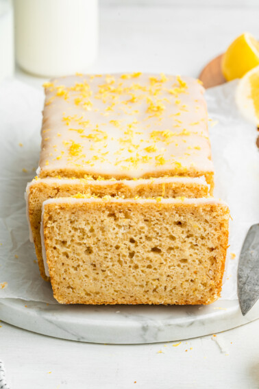 A lemon loaf topped with a lemon glaze and lemon zest. The loaf is cut so you can see the inside of the loaf.