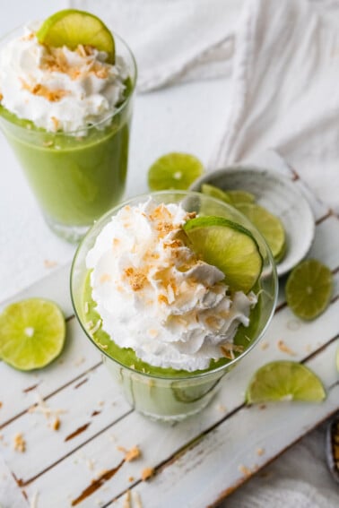 Two key lime pie smoothies in glasses topped with whip cream, toasted coconut and lime slices.