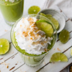 Two key lime pie smoothies in glasses topped with whip cream, toasted coconut and lime slices.