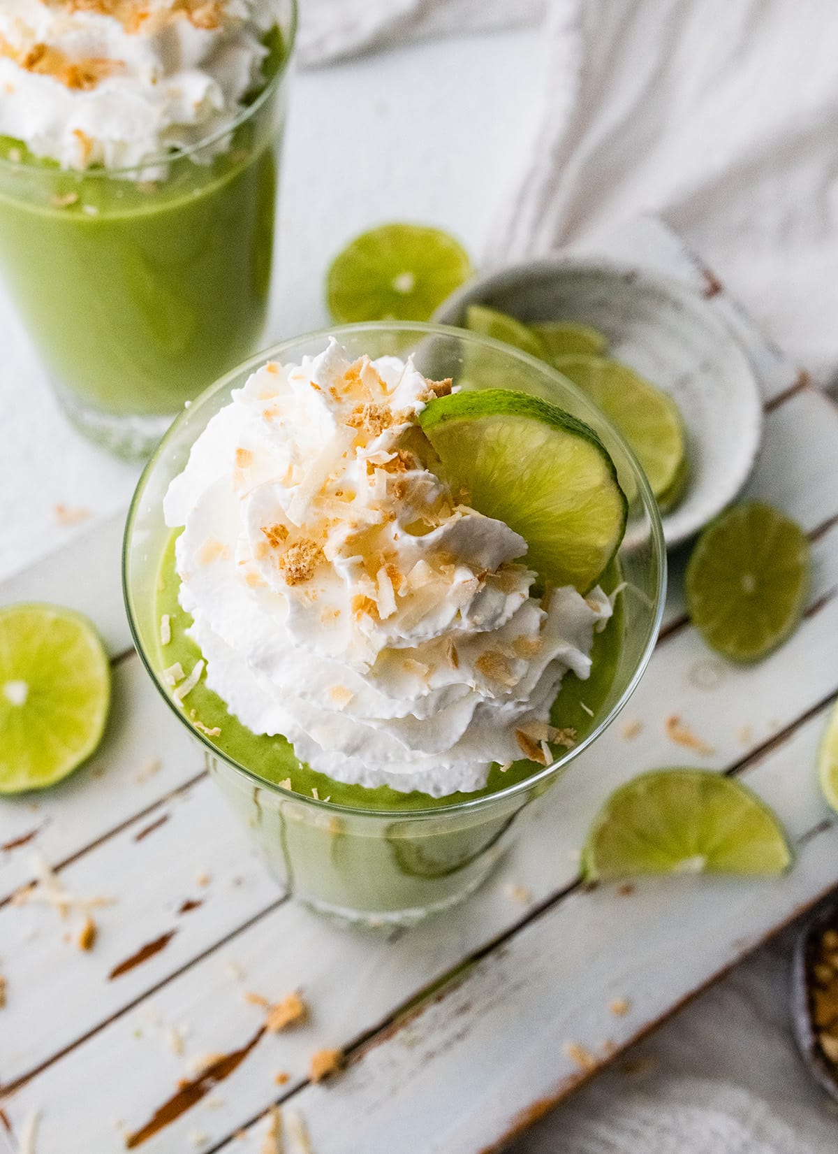 Two key lime pie smoothies in glasses topped with whip cream, toasted coconut and lime slices.