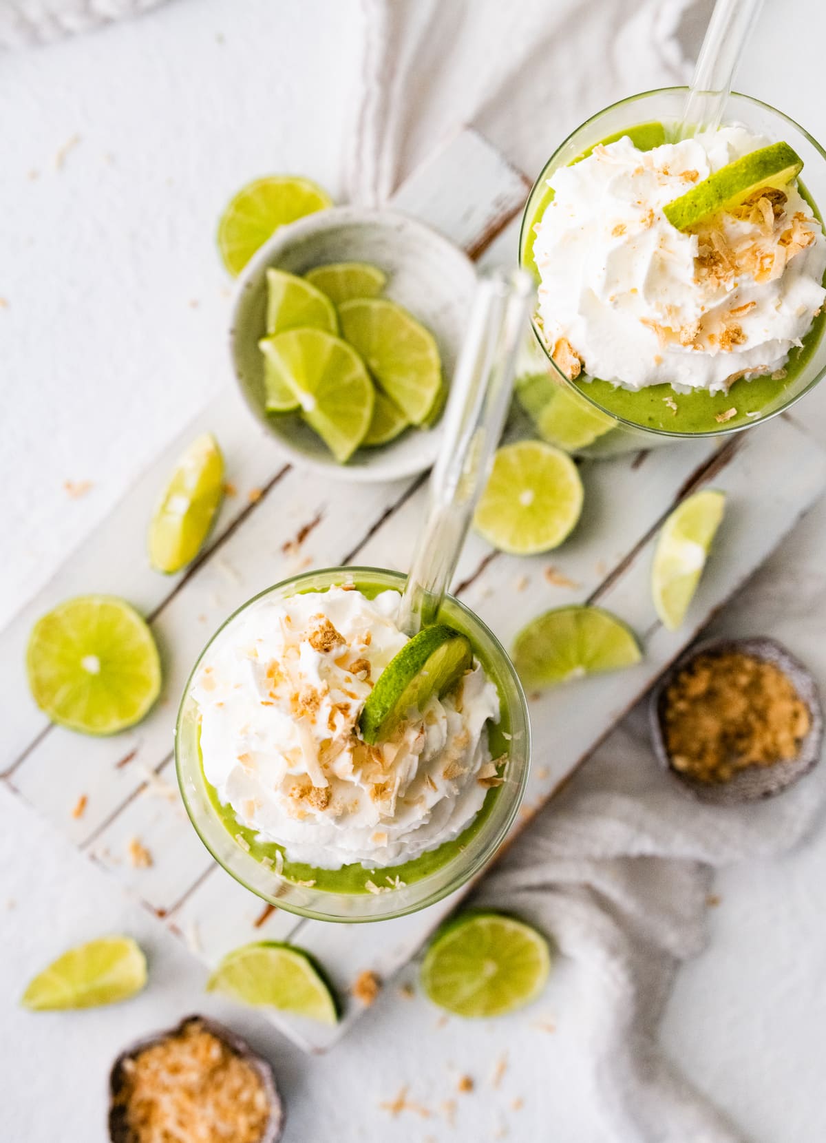 Overhead view of two key lime pie smoothies with straws, topped with whip cream, toasted coconut and lime slices.