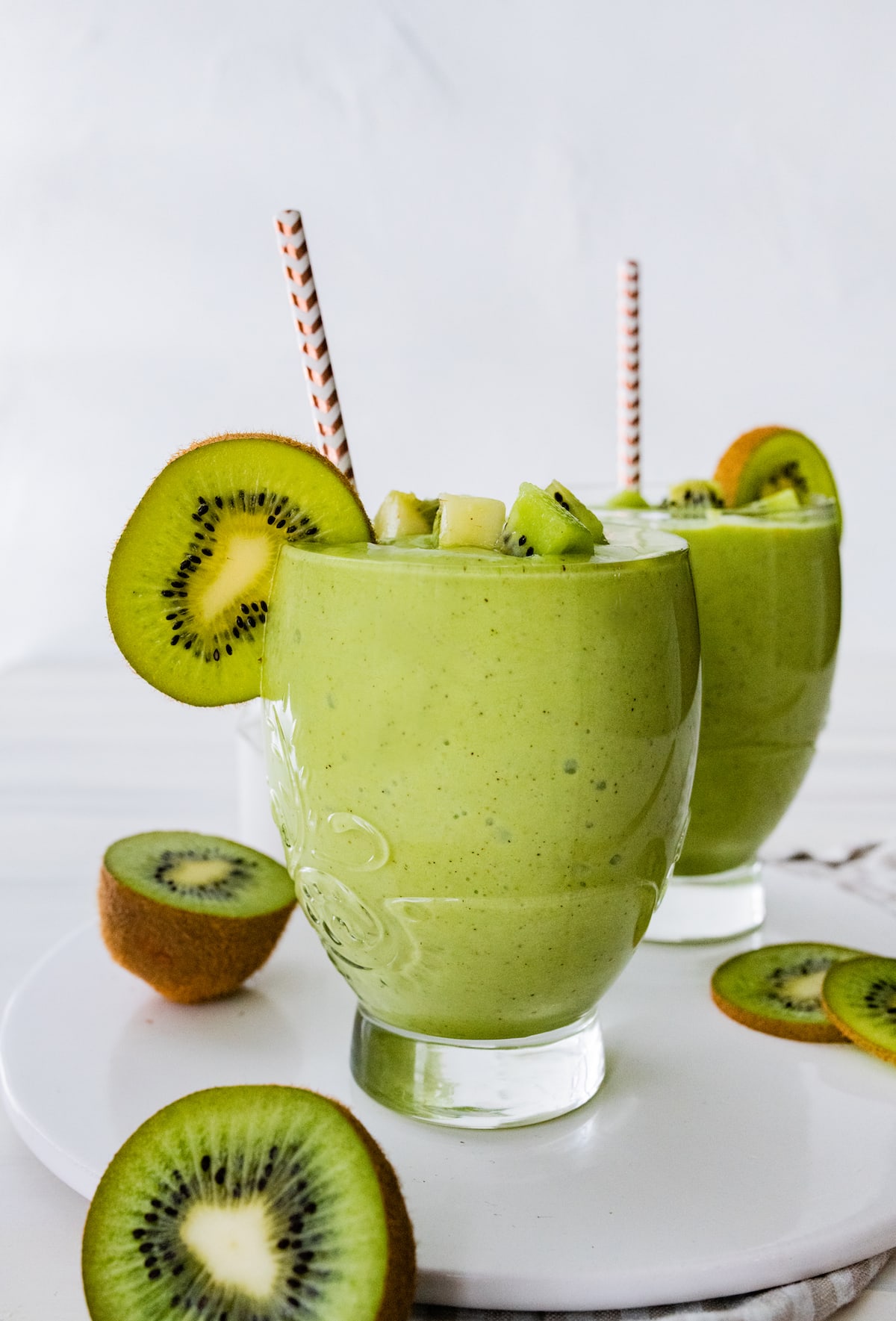 Two kiwi smoothies with straws and kiwi slices on side of glass.