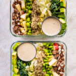 Two glass storage containers layered with detox salad ingredients: almonds, apple, edamame, quinoa, cabbage, kale and avocado. A small container of dressing is also in each of the containers.