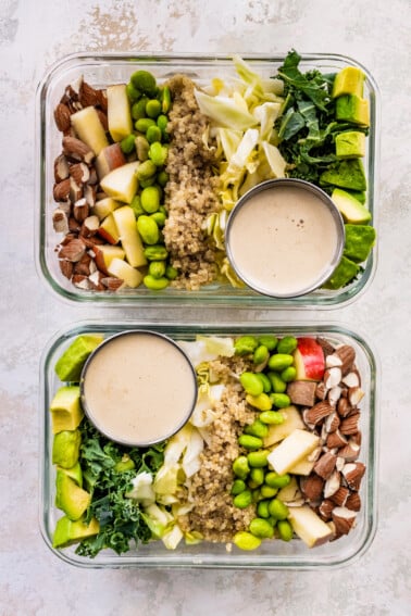 Two glass storage containers layered with detox salad ingredients: almonds, apple, edamame, quinoa, cabbage, kale and avocado. A small container of dressing is also in each of the containers.