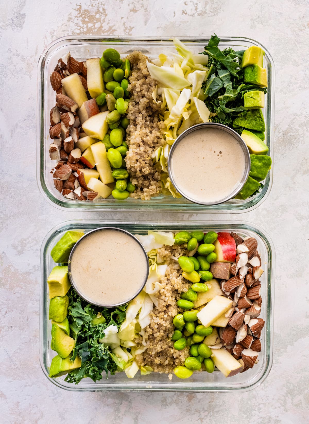 Two glass storage containers layered with almonds, apple, edamame, quinoa, cabbage, kale and avocado. A small container of dressing is also in the container.