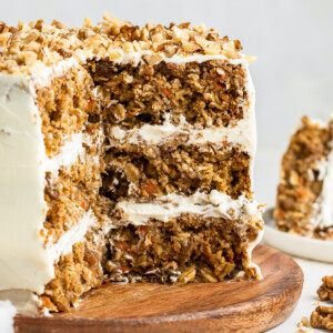 3-layer oatmeal carrot cake, frosted with chopped walnuts on top served on a wooden board. A few slices of the cake have been cut out exposing the inside of the cake.