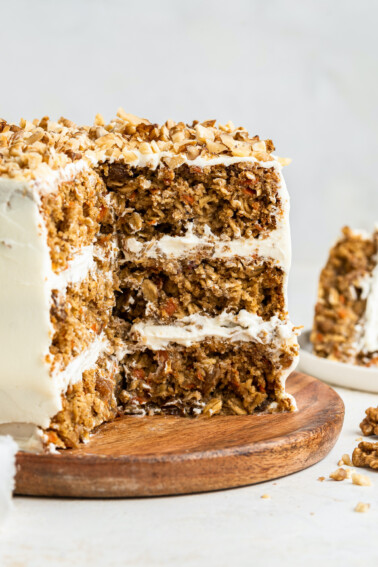 3-layer oatmeal carrot cake, frosted with chopped walnuts on top served on a wooden board. A few slices of the cake have been cut out exposing the inside of the cake.