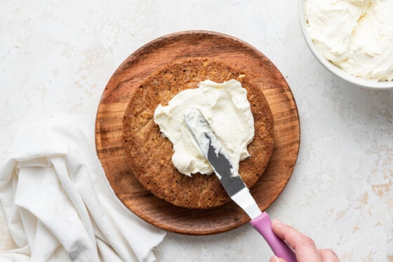 Woman's hand frosting one layer of the oatmeal carrot cake with a small cake spatula with a pink handle.