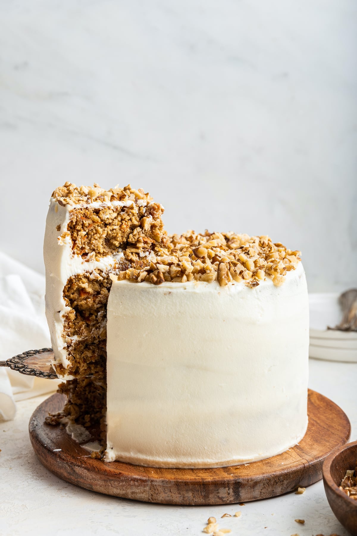 Oatmeal carrot cake on a wooden board with a slice being removed from it. The cake is frosted in a cream cheese frosting and has chopped walnuts on top.