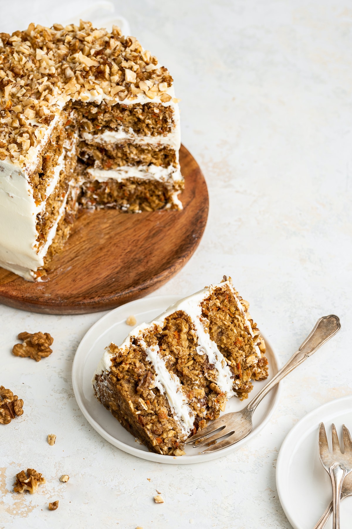 A slice of a 3-layer oatmeal carrot cake served on a white plate with a fork. The full 3-layer oatmeal carrot cake is behind the plate on a wooden board with the center exposed.