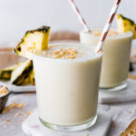 Two pineapple smoothies with straws, topped with toasted coconut and fresh pineapple slices.