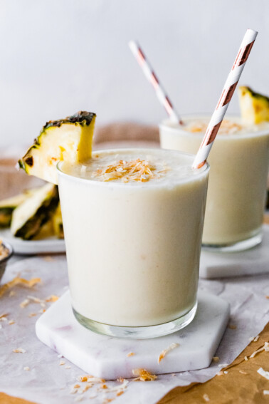 Two pineapple smoothies with straws, topped with toasted coconut and fresh pineapple slices.