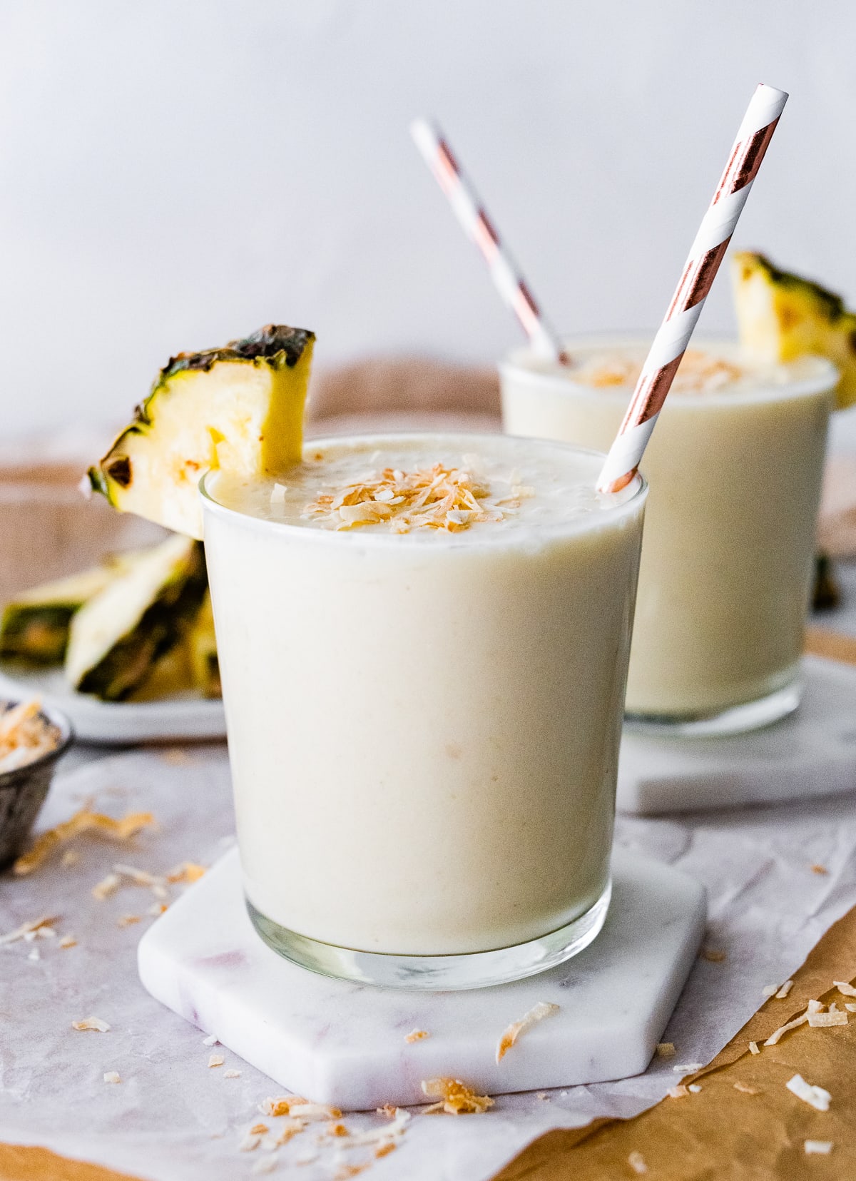 Two pineapple smoothies with straws, topped with toasted coconut and fresh pineapple slices.