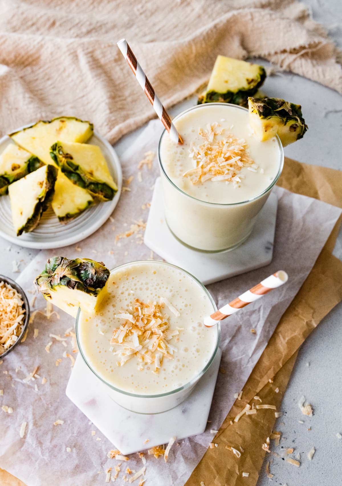Two pineapple smoothies with straws, topped with toasted coconut and fresh pineapple slices.