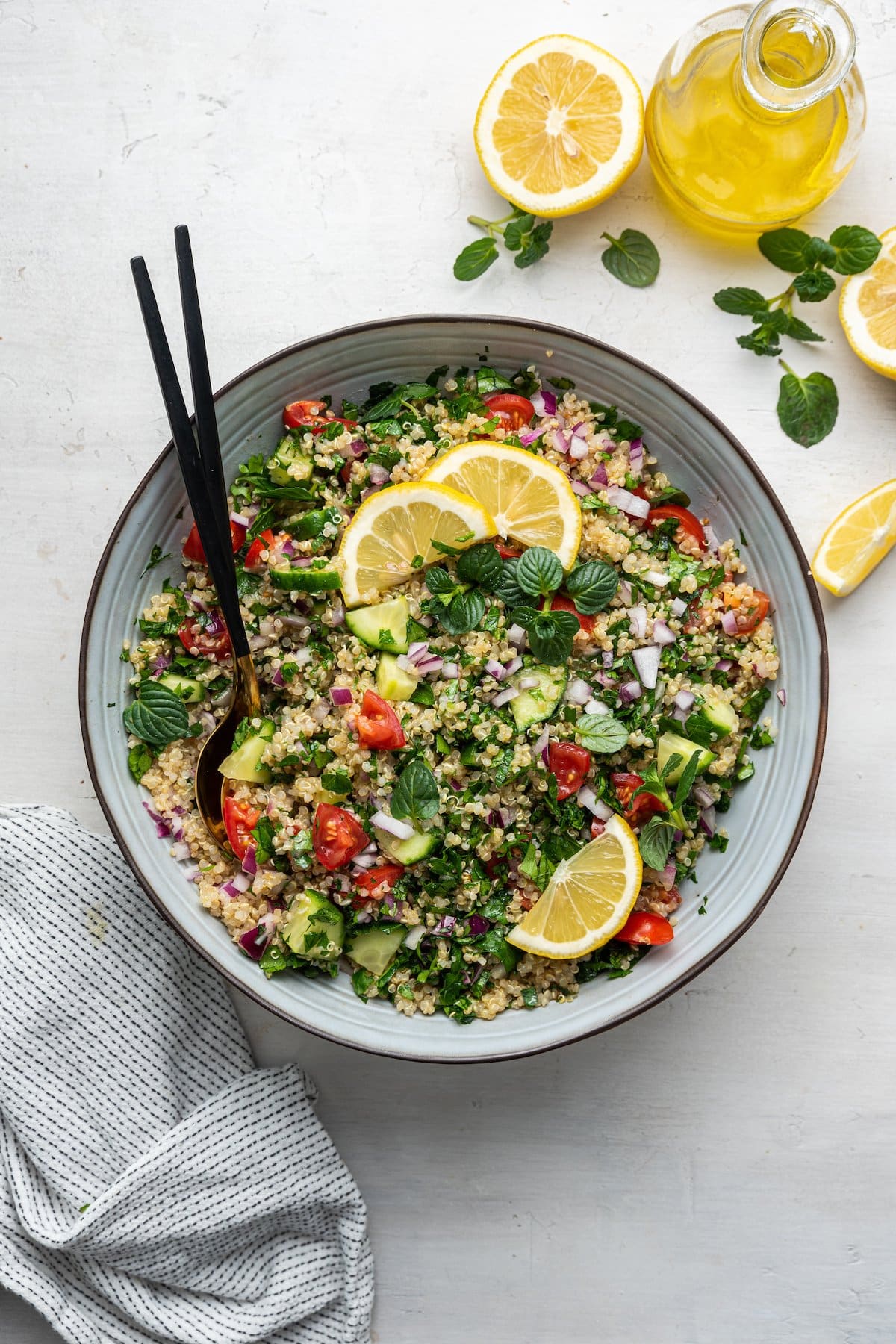 Quinoa tabbouleh in a serving bowl with serving spoons topped with fresh lemon slices and fresh herbs.