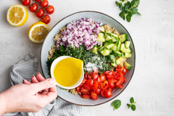 Woman's hand pouring olive oil over quinoa tabbouleh ingredients in a serving bowl.