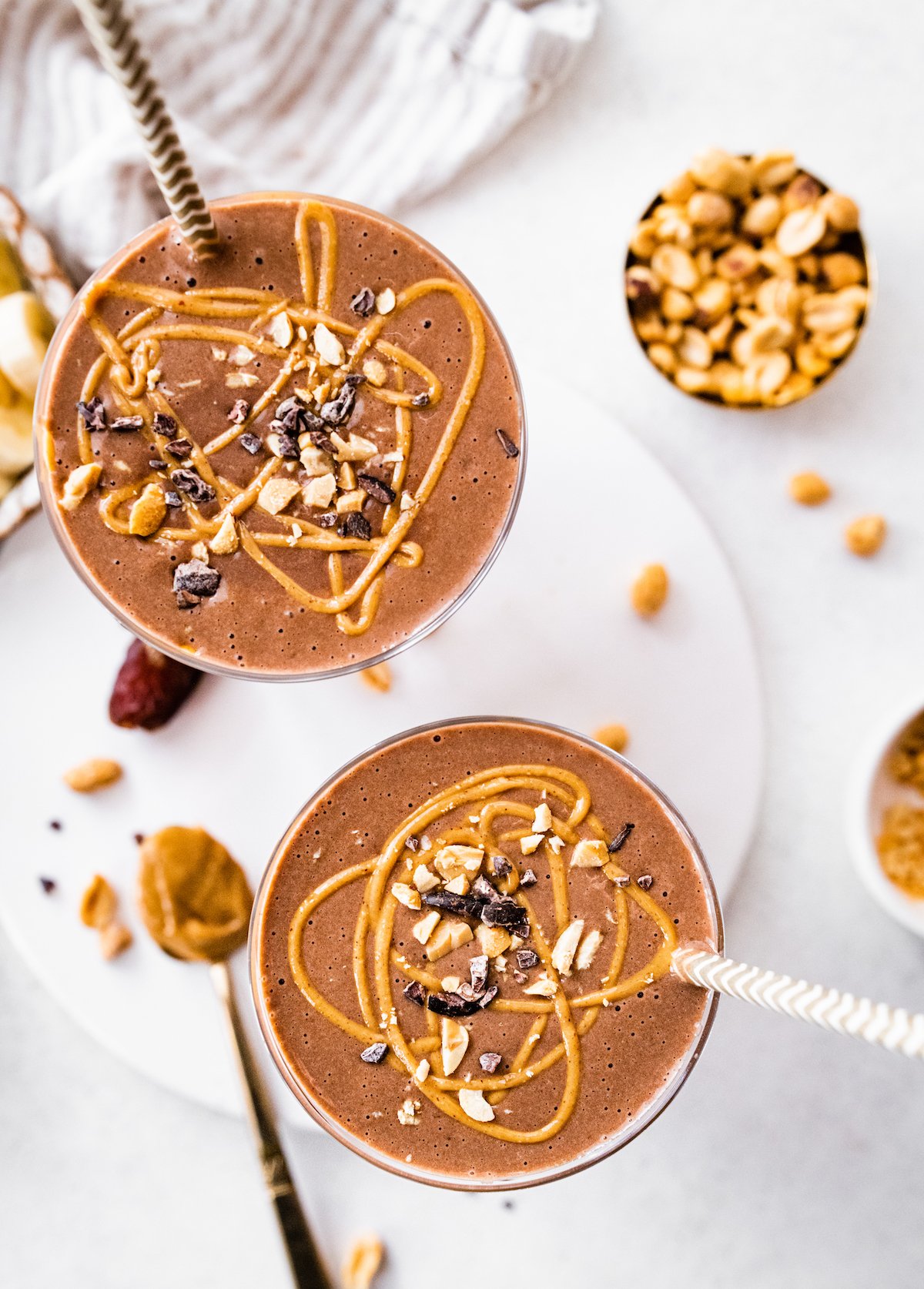 Two close up snickers protein smoothies with straws, topped with cacao nibs, chopped peanuts and a drizzle of peanut butter.