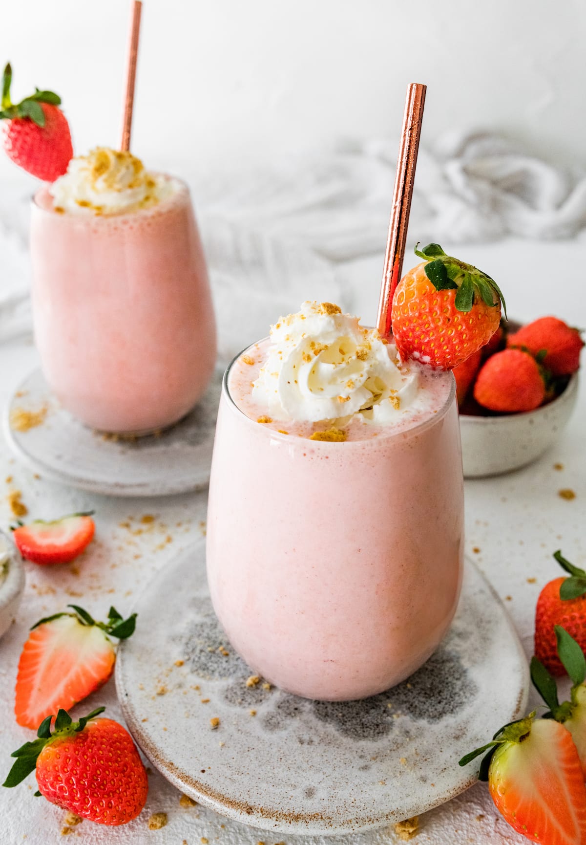 Two strawberry cheesecake smoothies with straws, topped with whipped cream, graham crackers and fresh strawberries.
