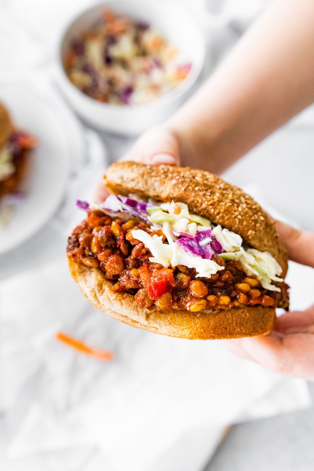 Woman's hand holding a vegan Sloppy Joe on a whole wheat bun topped with coleslaw.