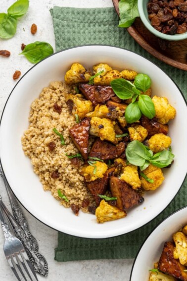 A white bowl containing the curried cauliflower and tempeh over a bed of quinoa and garnished with fresh basil leaves.