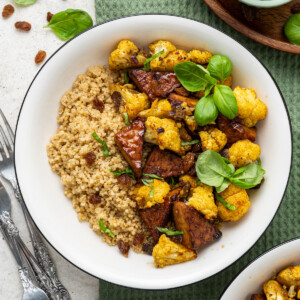 A white bowl containing the curried cauliflower and tempeh over a bed of quinoa and garnished with fresh basil leaves.