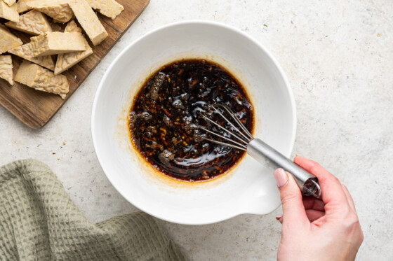 A woman's hand holding a whisk to whisk the marinade for the tempeh. The marinade includes maple syrup, balsamic vinegar, tamari, oil, and minced garlic.