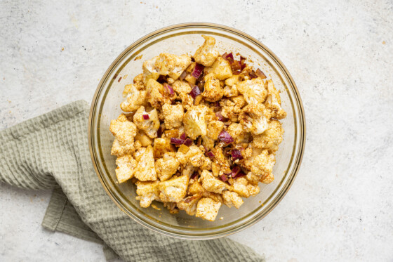 A medium glass bowl containing cauliflower florets with chopped red onion and many spices.