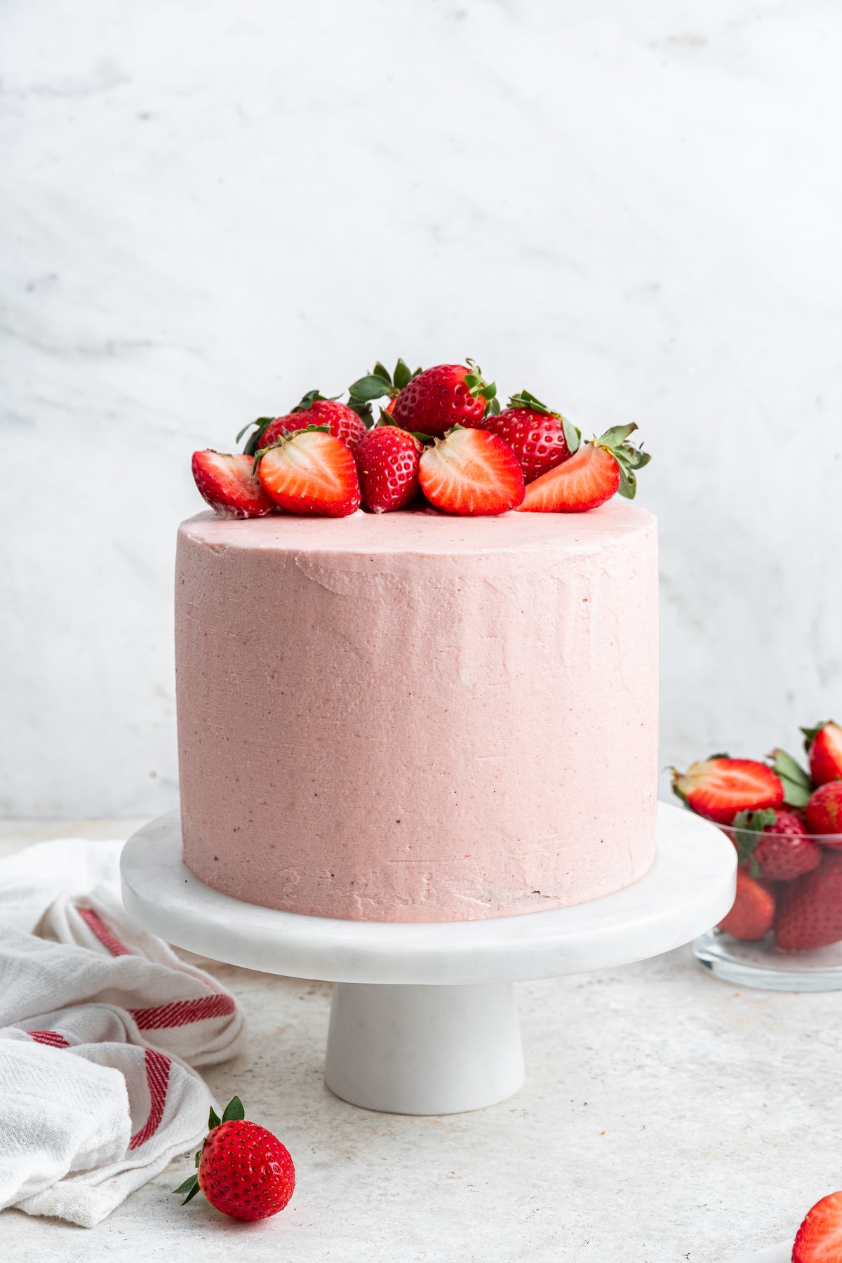 A strawberry cake with pink frosting and fresh strawberries on top on a small white pedestal.