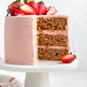 A strawberry cake with pink frosting sits on a small white pedestal with fresh strawberries on top and one slice cut out.