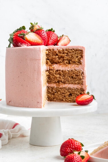 A strawberry cake with pink frosting sits on a small white pedestal with fresh strawberries on top and one slice cut out.