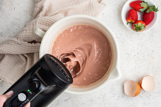 A woman's hand holding a hand-held mixer, mixing the dry and wet ingredients of the strawberry cake filling in a white bowl.