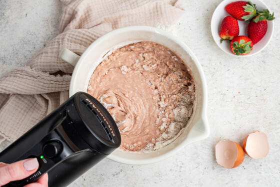 A woman's hand holding a hand-held mixer, mixing the dry and wet ingredients of the strawberry cake filling in a white bowl.