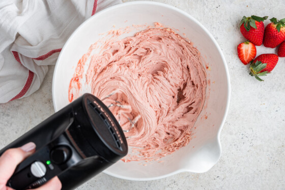 A woman's hand holding a hand-held mixer while mixing the strawberry cake frosting in a large white bowl.