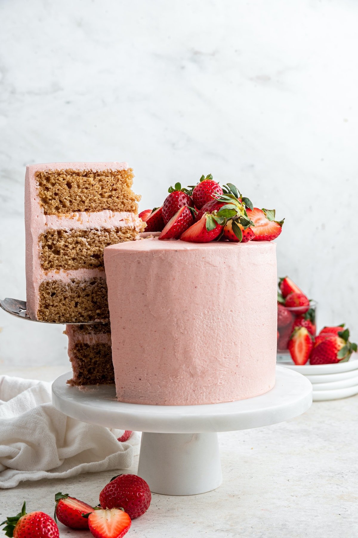 A slice of strawberry cake is being served off a small pedestal where the cake stands. The cake is covered in pink frosting and topped with fresh strawberries.