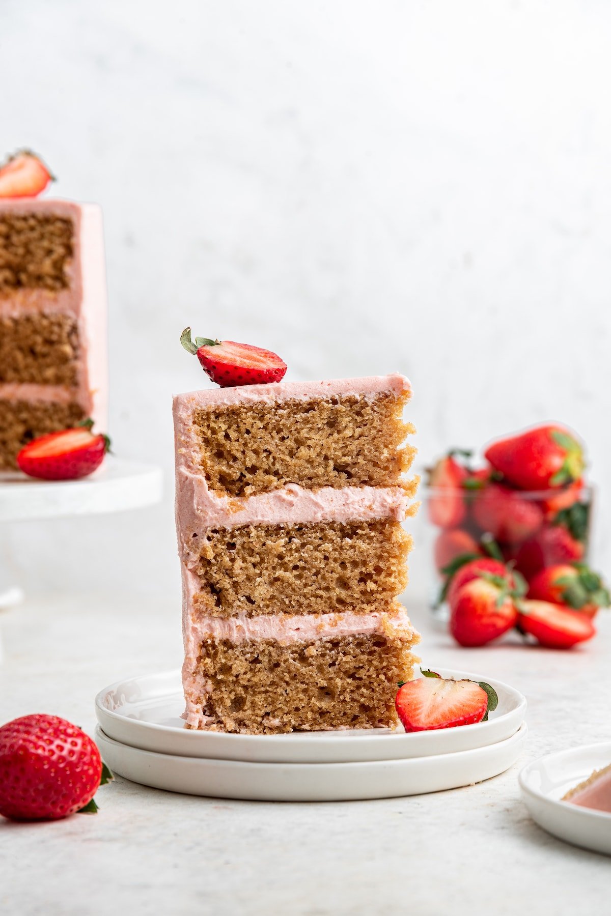 Two small white plates stacked on each other hold a slice of strawberry cake standing vertically with a glass container full of strawberries in the background.