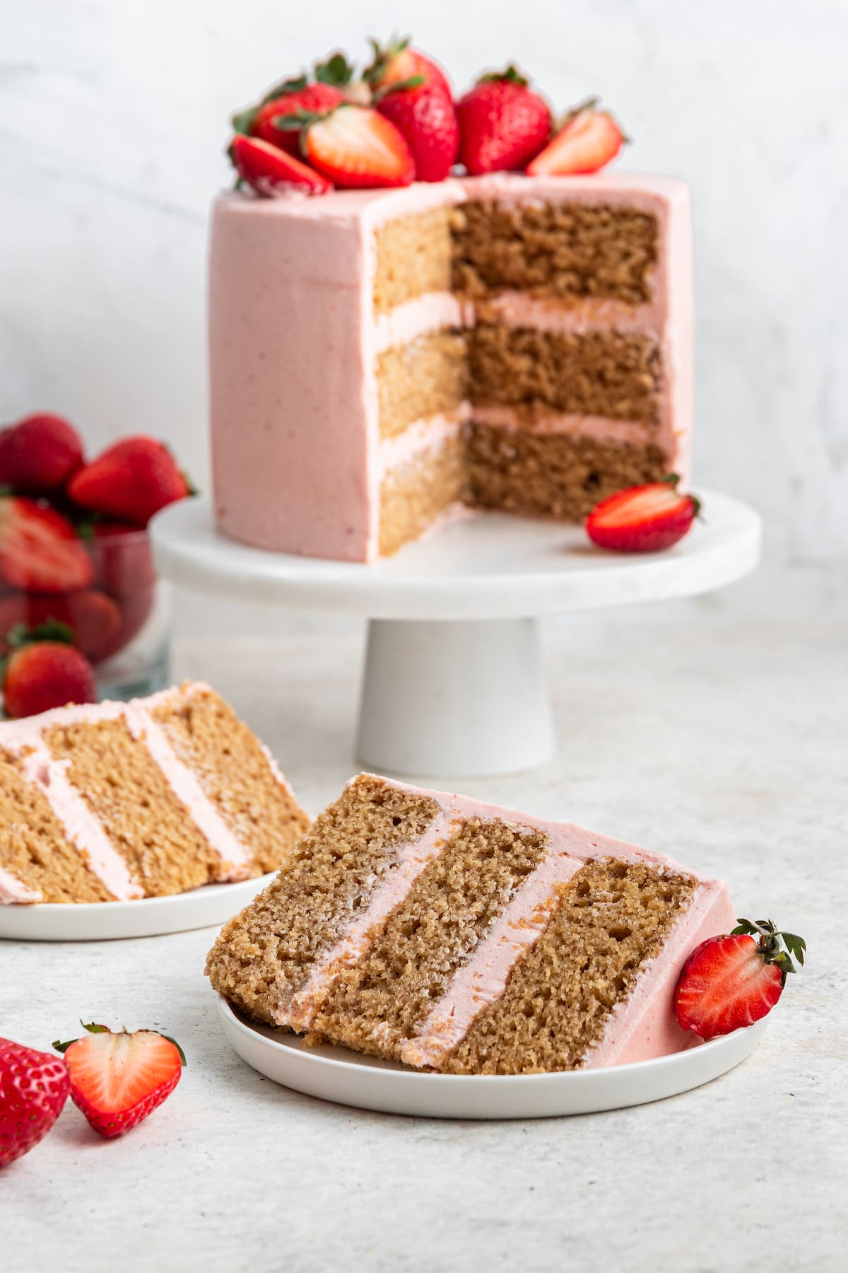 Two slices of strawberry cake on separate small white plates. The strawberry cake is on a small pedestal in the background with pink frosting and fresh strawberries on top of the cake.