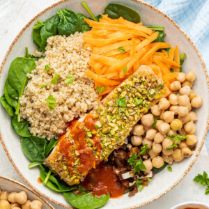 A pistachio salmon salad served in a white bowl. The bowl contains a baked salmon filet, chickpeas, shredded carrots, quinoa, chopped dates, spinach, and spicy harissa dressing.