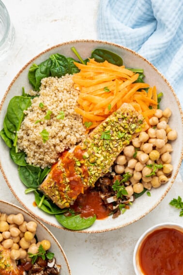 A pistachio salmon salad served in a white bowl. The bowl contains a baked salmon filet, chickpeas, shredded carrots, quinoa, chopped dates, spinach, and spicy harissa dressing.