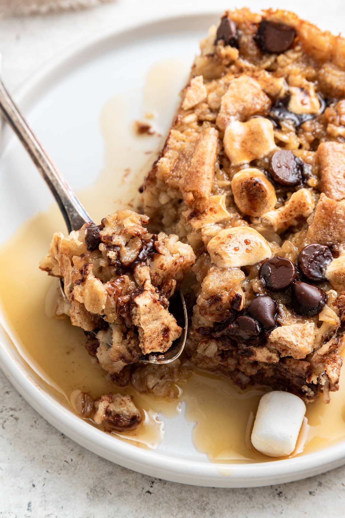 A close-up of a slice of the s'mores baked oatmeal lightly covered in maple syrup on a white plate. A metal spoon holds a small piece of the baked oatmeal.