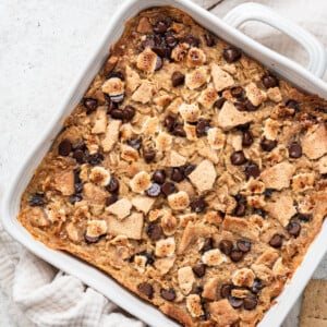 A white square baking dish with the s'mores baked oatmeal inside after baking in the oven. The top is lightly browned with chocolate chips, mini marshmallows, and crushed graham crackers.