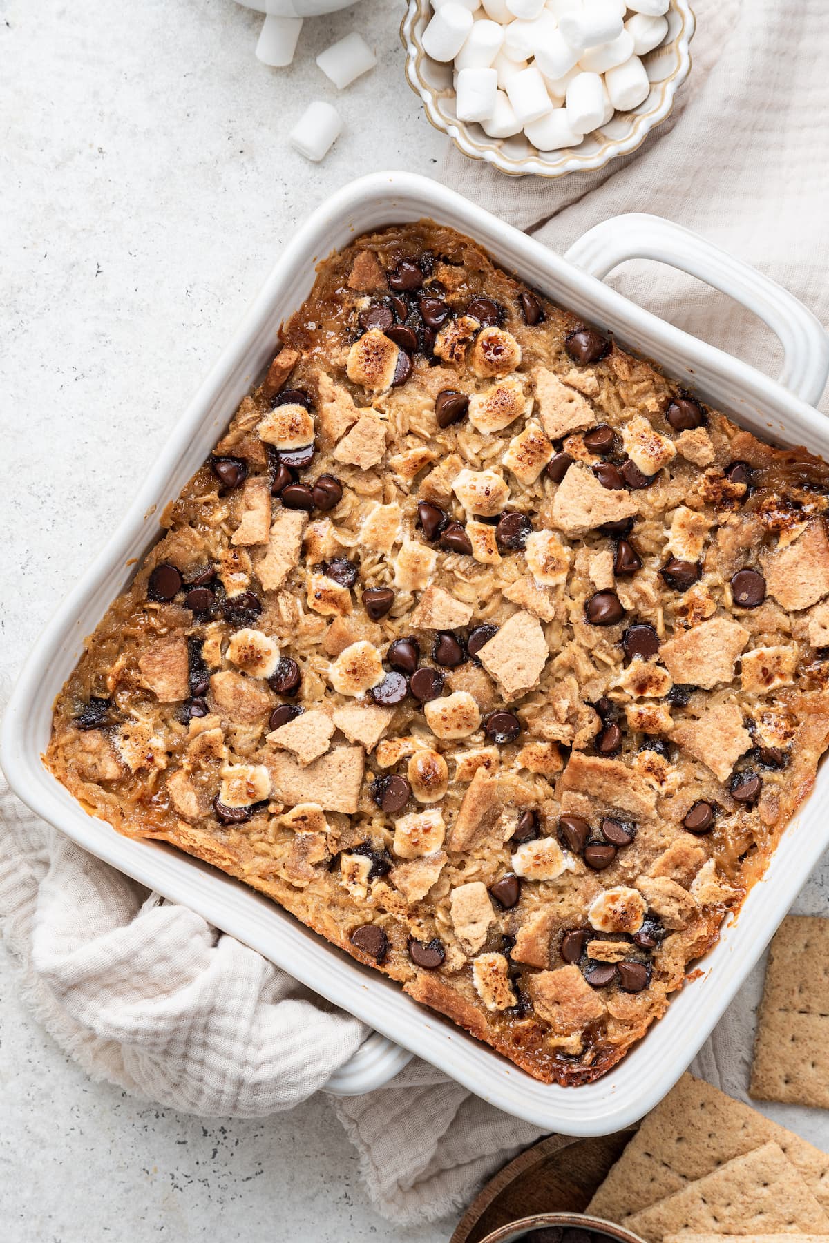 A white square baking dish with the s'mores baked oatmeal inside after baking in the oven. The top is lightly browned with chocolate chips, mini marshmallows, and crushed graham crackers.